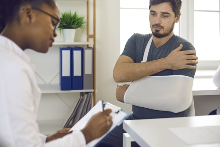 Man in sling with injured arm speaks with medical professional in a brightly lit office
