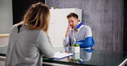 Man with broken arm and neck brace sits at a desk with a personal injury attorney to discuss his case